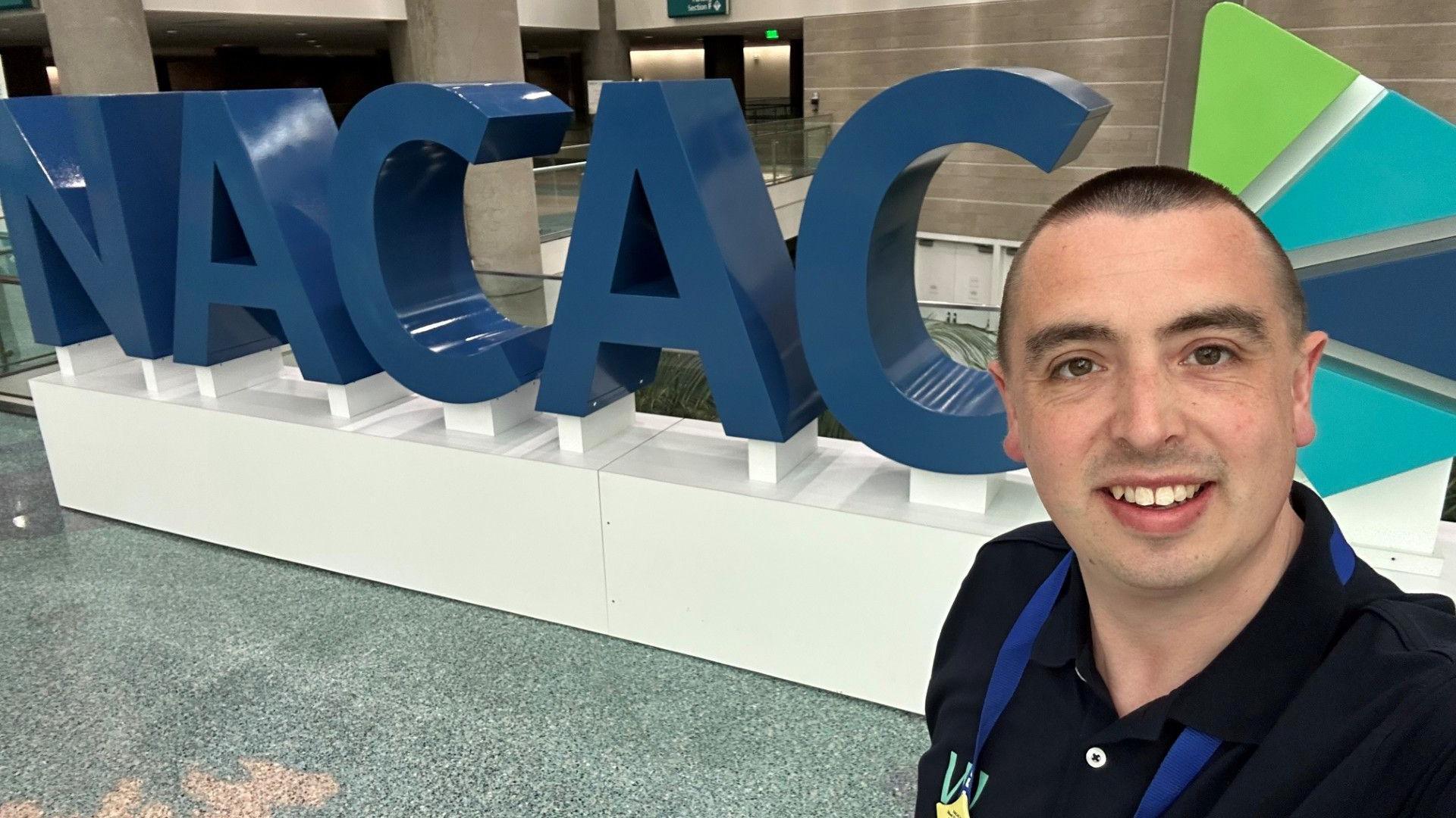 A white man in his 30s with a buzz cut smiles in a selfie taken in front of large 3D letters which spell out the acronym NACAC. He is wearing a dark polo shirt and a lanyard.