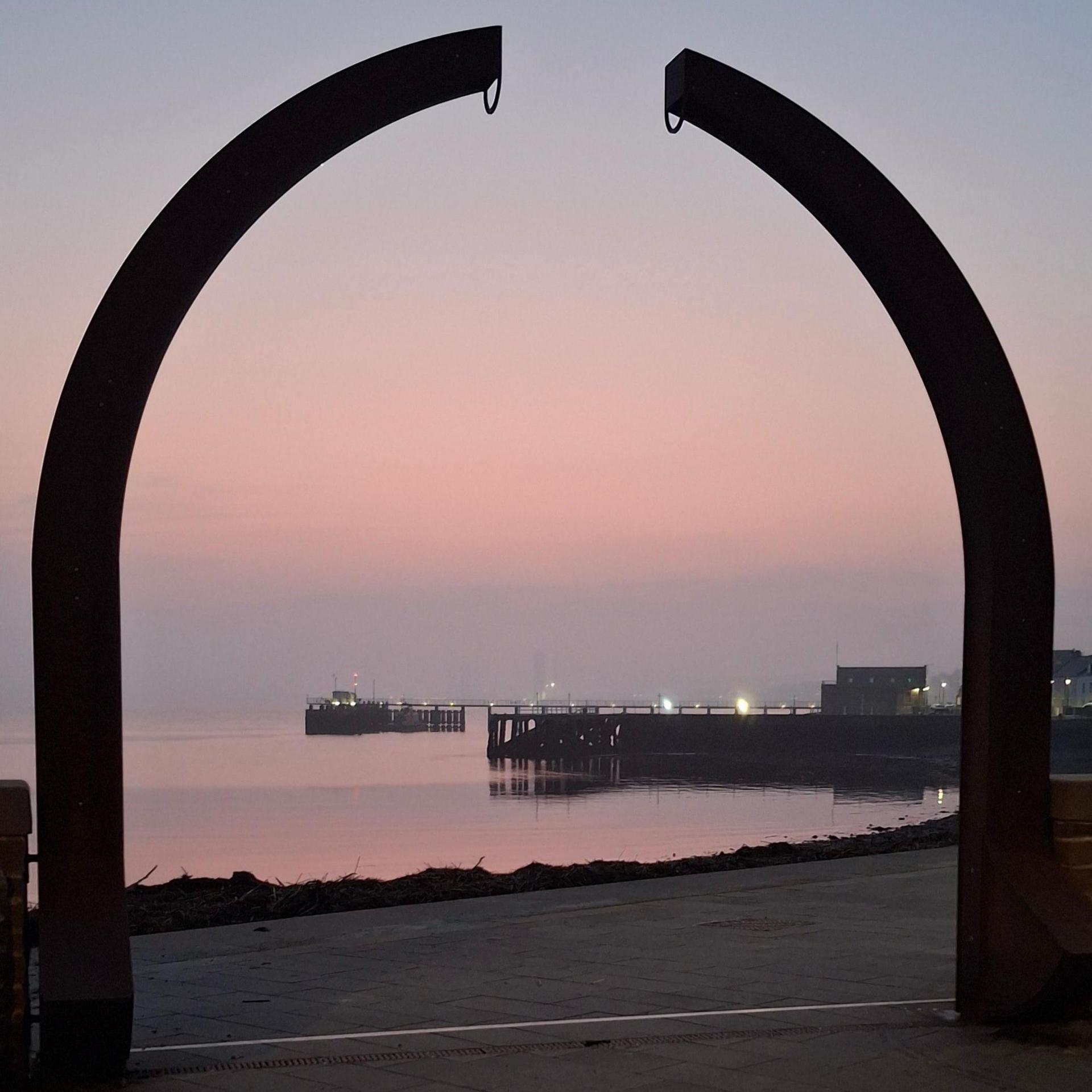 A shot of the River Tay through an archway. The sky is pink and blue with touches of orange. A pier can be seen in the background. It is dark and shadowed against the lighter background. The archway is also dark and extends about three-quarters of the way up the image.