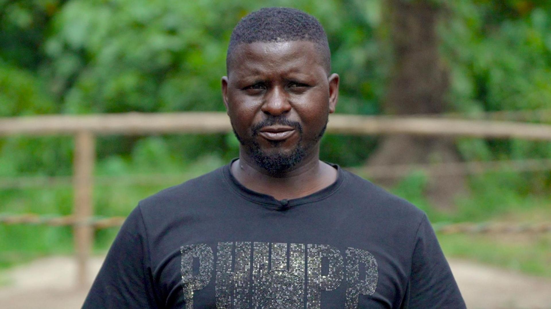 Daniel Bumba wears a black T-shirt and looks at the camera as a wrestling ring is seen out of focus in the background behind him