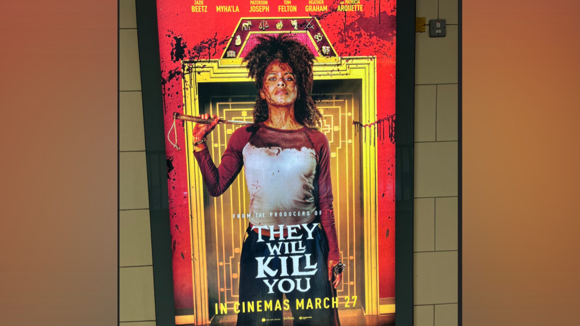 A film poster on an underground station wall shows a blood-splattered woman standing in front of an ornate doorway. She wears a blood-stained shirt, has blood splattered across her face, and holds a weapon resting over her shoulder. Red blood splatters cover the background behind her. The text reads: