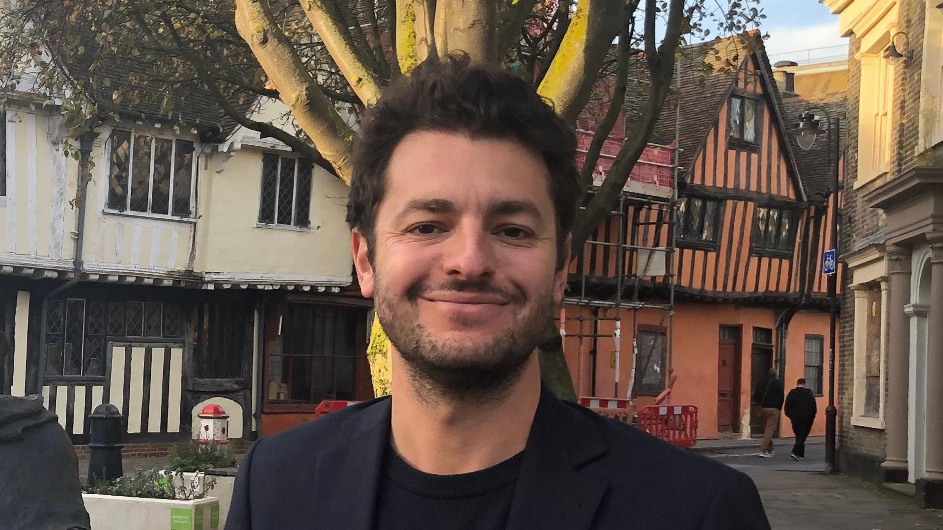 A photo of Ipswich MP Jack Abbott. He is facing the camera and smiling. He has dark hair and a dark beard. Tudor-style buildings can be seen behind him while pedestrians walk nearby.