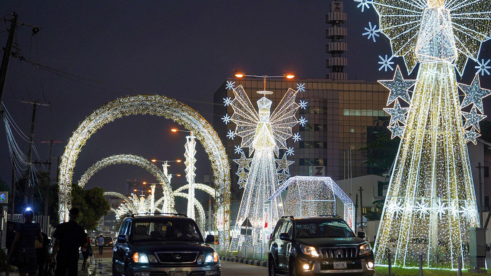 Cars drive past tall bright cone shaped lights with geometric angels with more lights arching across the road in the background