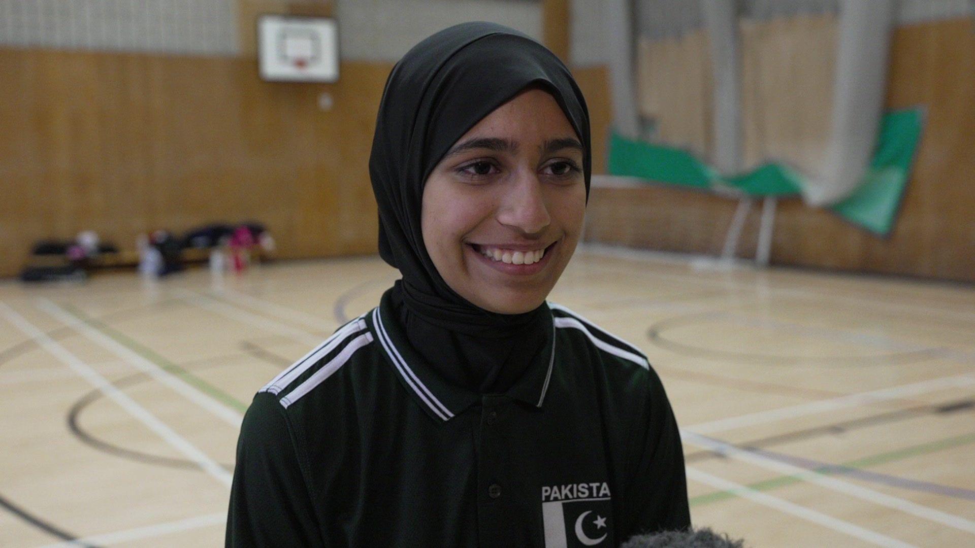 Haleema smiles widely to the left of the camera. she is wearing a black hijab and a black polo shirt with the Pakistan team logo top left. she is standing in an indoor netball court with the floor markings and netting and a hoop on the wall visible in the blurred background behind her