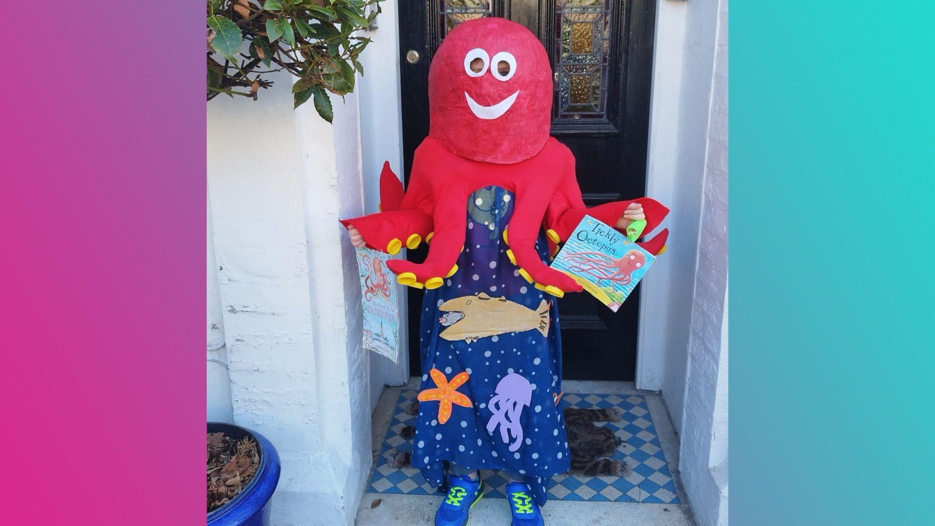 A boy wearing a red cape with eight octopus tentacles and a red paper mache head with round eyes and a big smile. Underneath the cape, the boy is wearing a navy blue tunic that's been made to look like the sea, covered in fish, jellyfish and starfish.