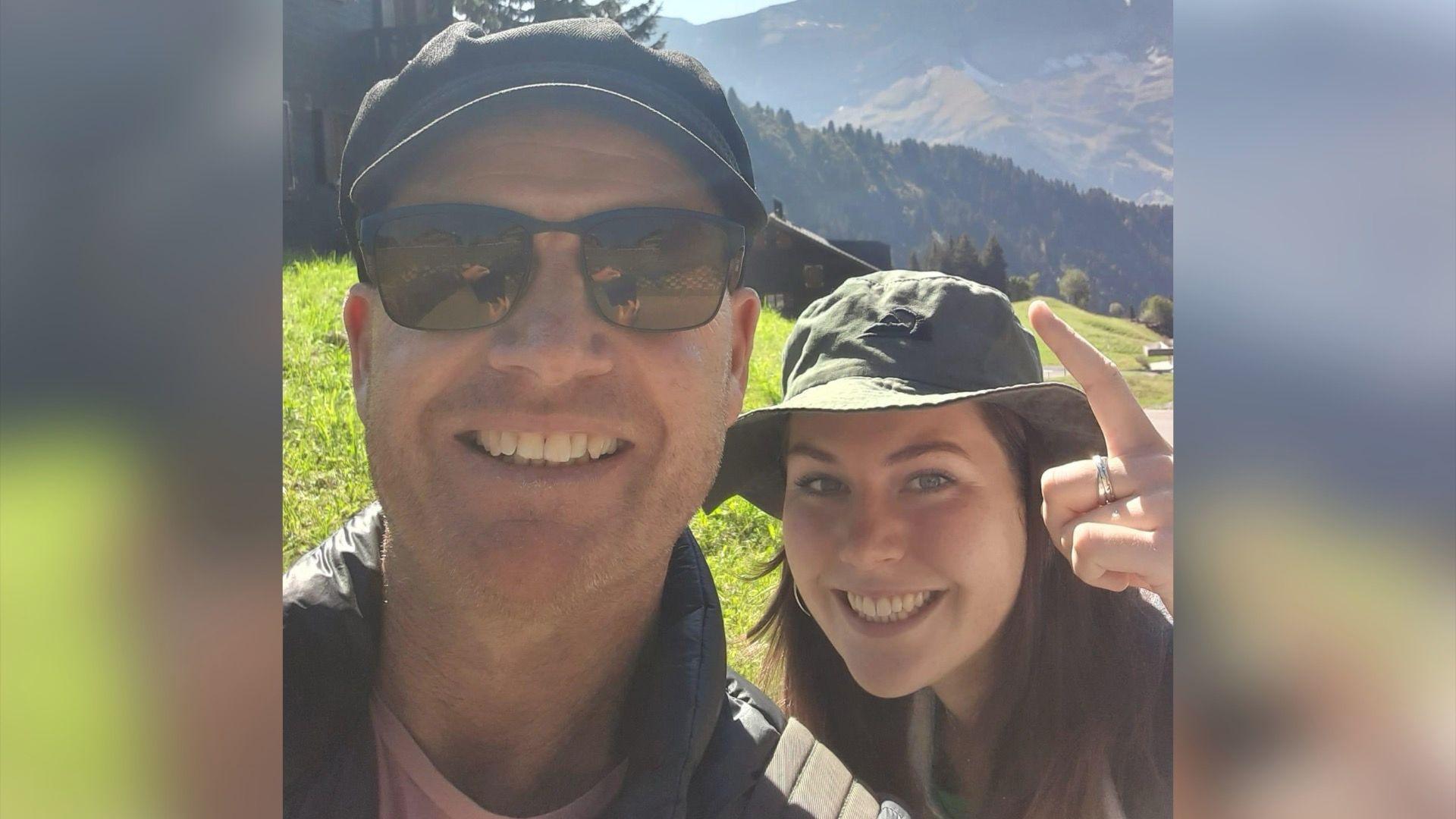 Man in hat on left smiling with woman behind also smiling in a hat in front of a forest view