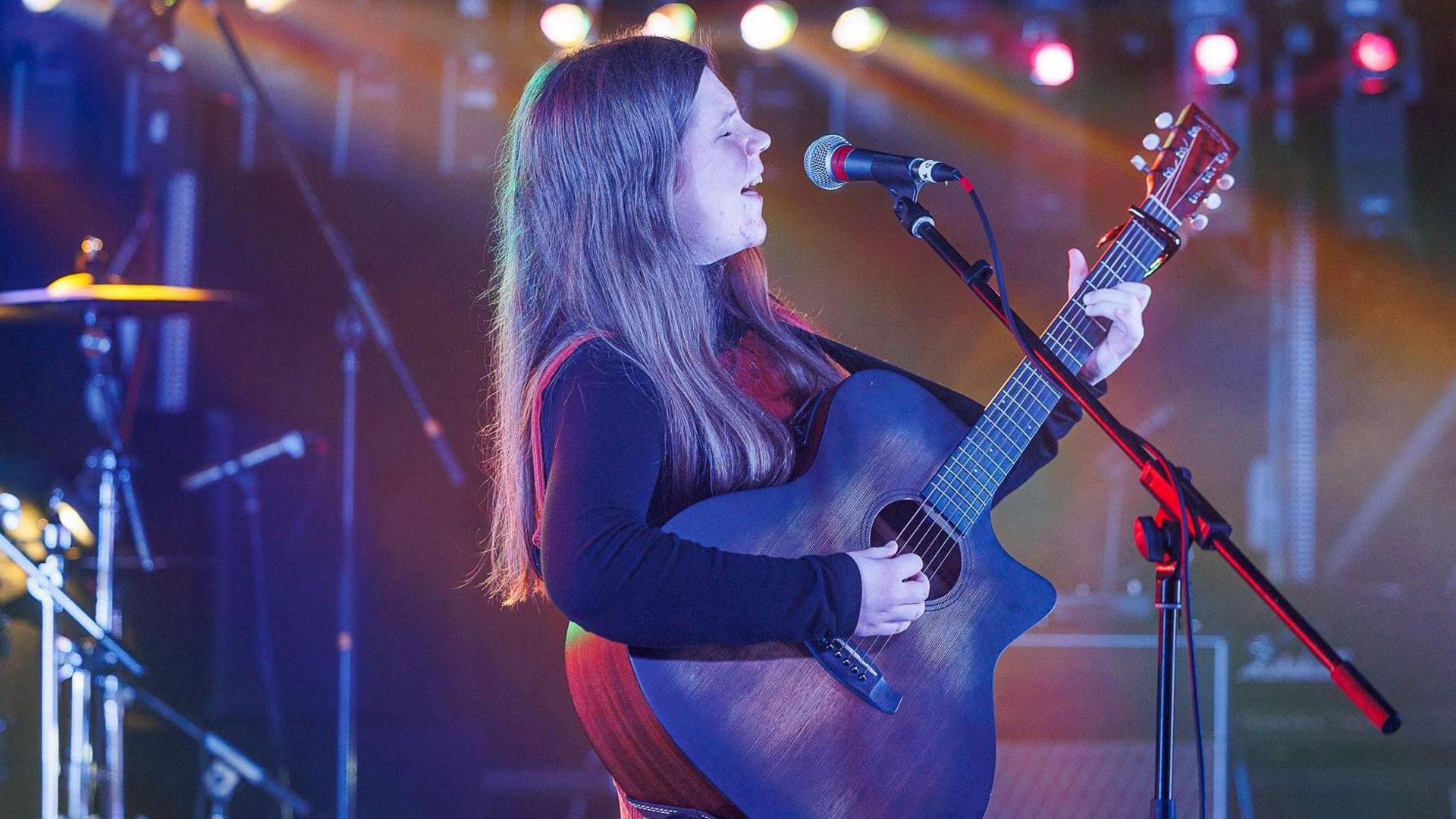 Isla Scott, who is wearing a blue long sleeved top, is standing side on to the camera as she stands playing a guitar and singing into a microphone. There are stage lights in the background.