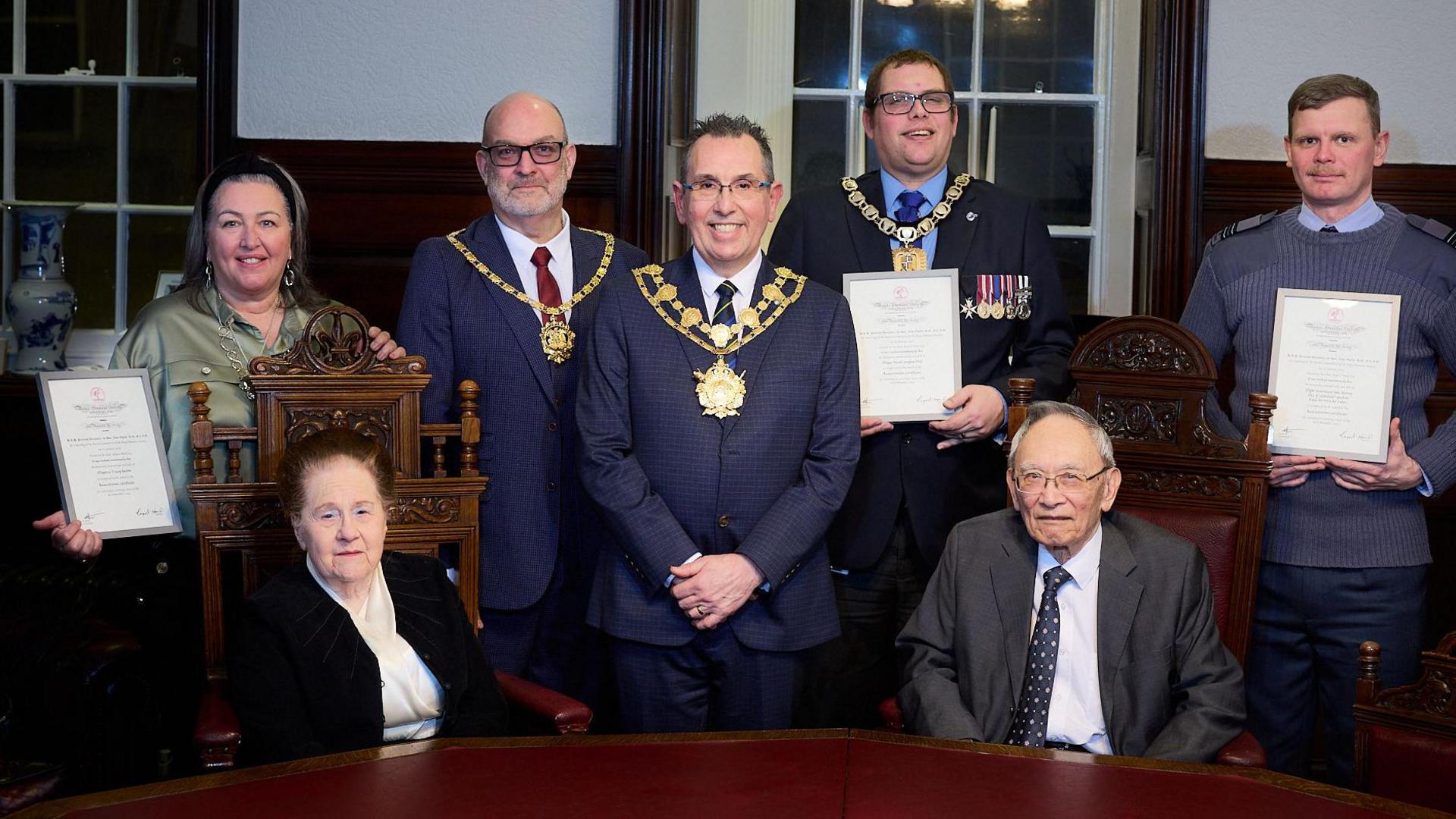 Sat in front of a round table are an older couple, Diana and Rudy Foo. Behind them are the mayor and mayoress who saved Rudy's life, and an officer from the Royal Air Force Air Cadets. They are all holding a certificate. In between them are three white men, all wearing mayoral chains, one is holding a certificate.