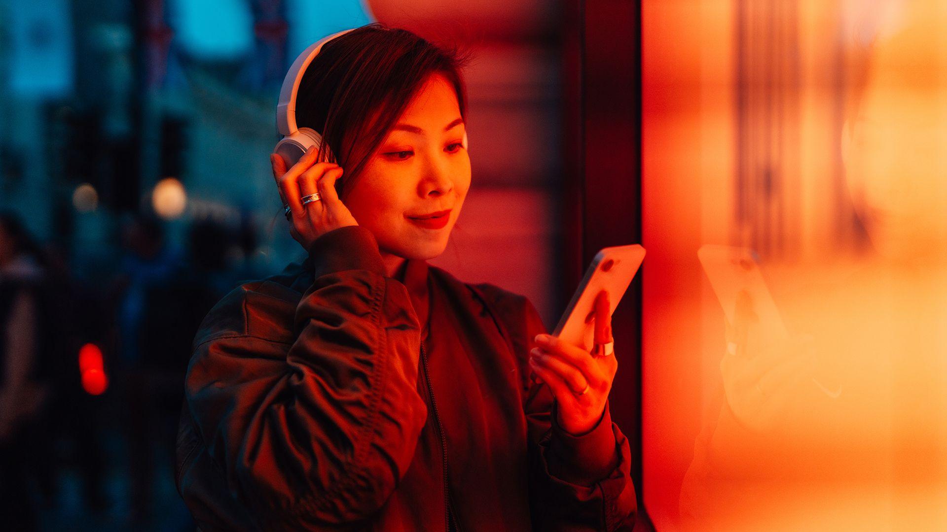 Woman listening to music on her smartphone, wearing wireless headphone while standing on London street in evening light.