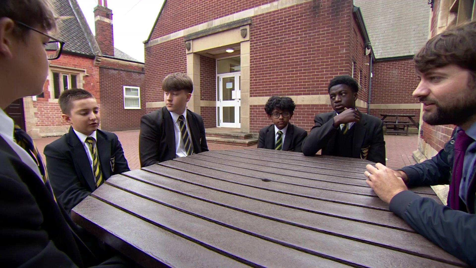 Five pupils sit at a table outside school buildings, a BBC reporter is seated at the same table on the right of frame.
The schoolboys are all wearing black blazers and yellow and black diagonally striped ties.