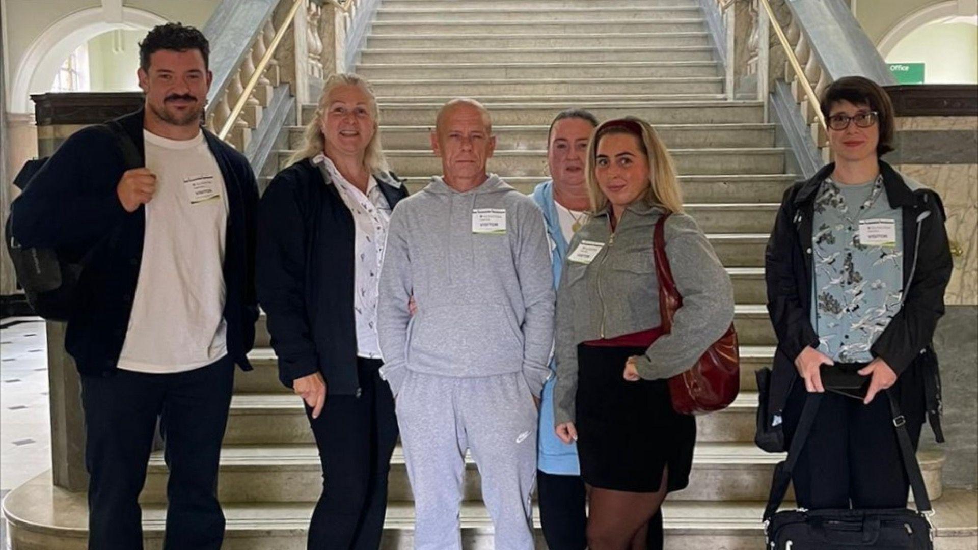 A group of six individuals standing in front of a grand staircase inside a historic building with marble walls and ornate railings. They are wear visitor badges.
