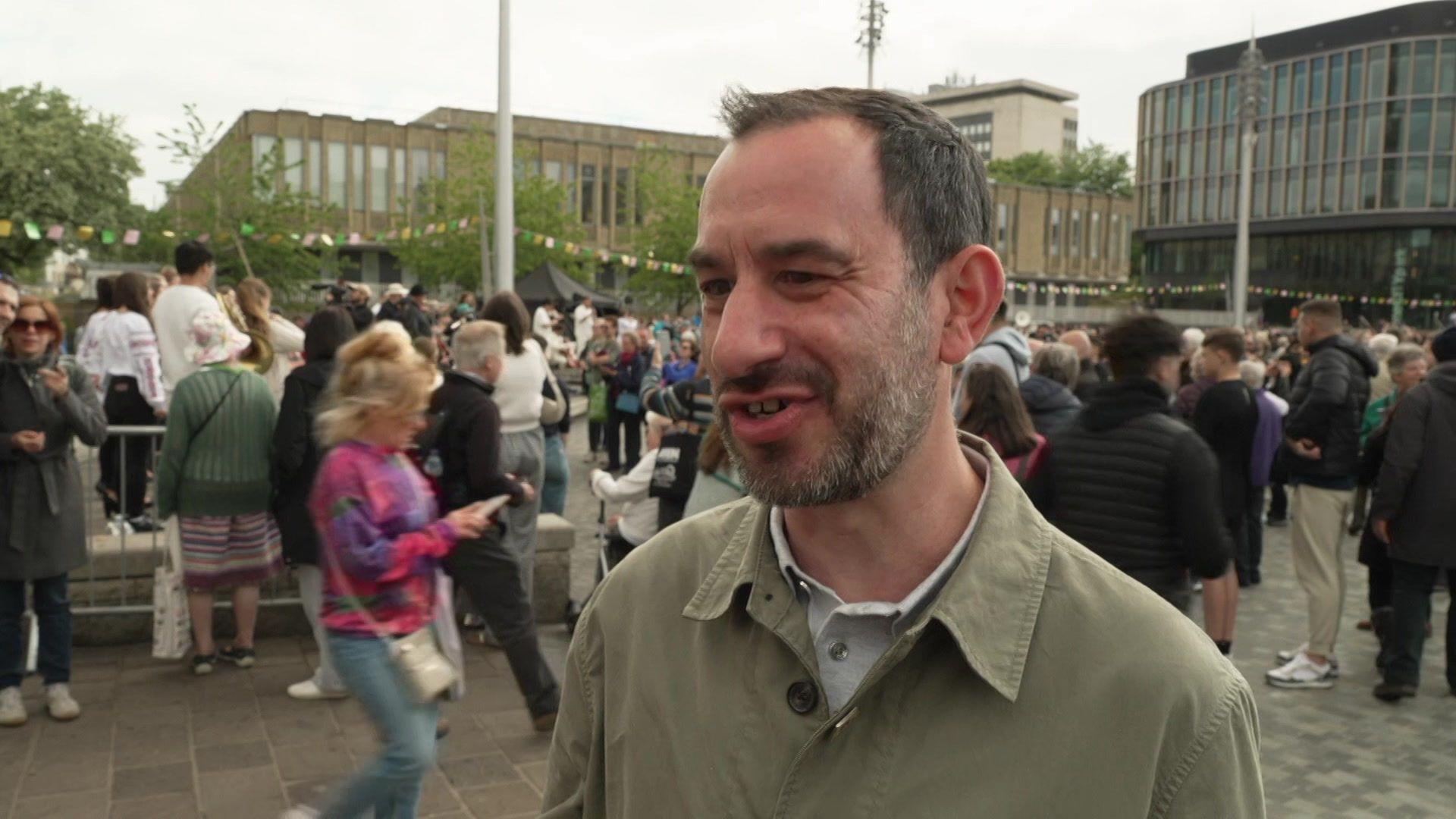Jonathan Harper speaking outside in Bradford. He is wearing an overshirt and has a short beard. There are lots of people gathered in the background.