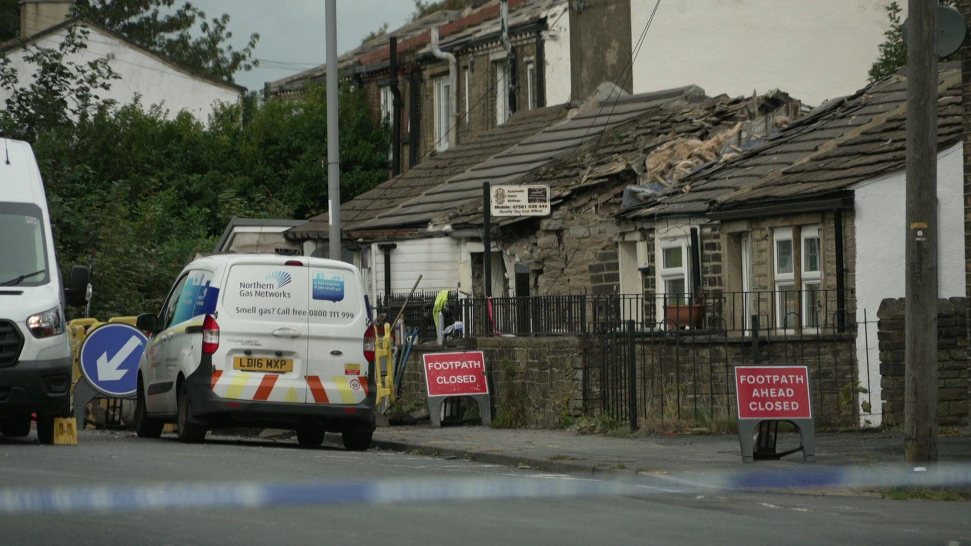 A Northern Gas Network van parked on a quiet street behind blue and white police tape. There are red 'footpath closed' signs on the pavement. A person in a yellow jacket can be seen in the distance working on rubble.