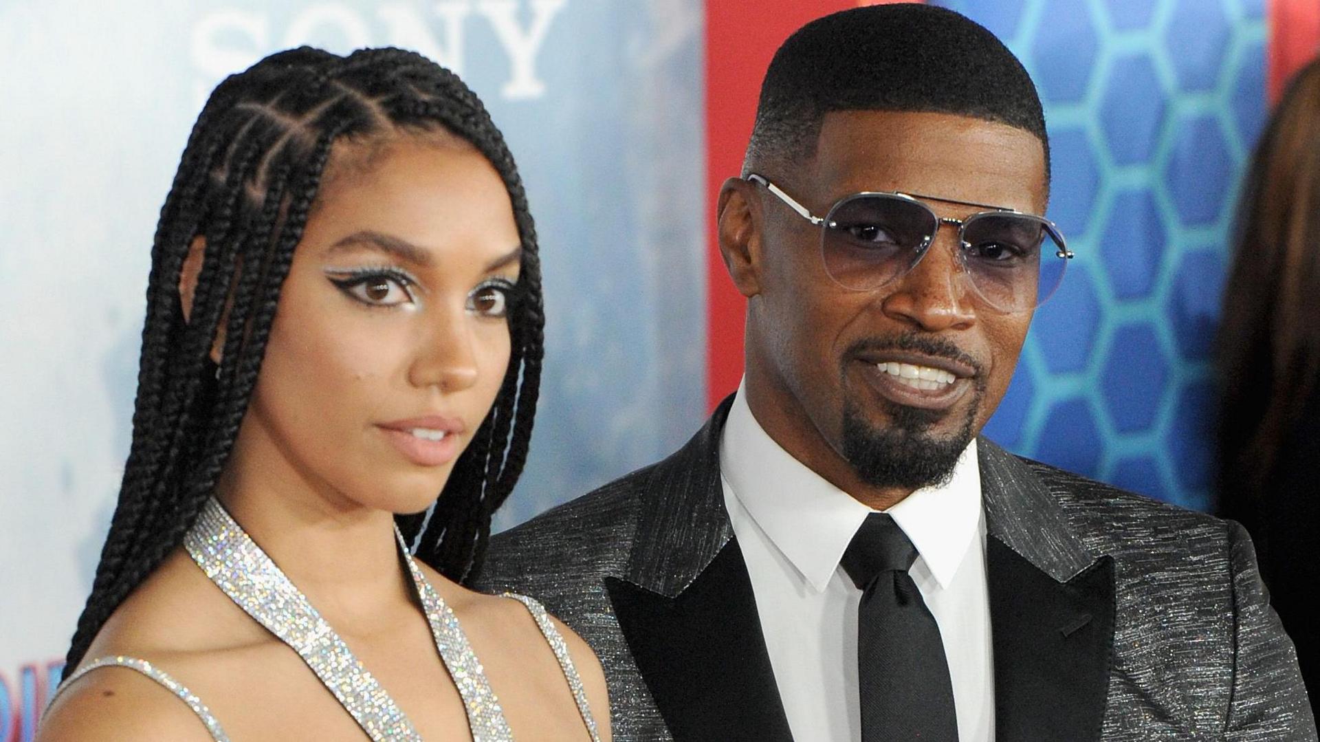 Corinne Foxx with her long hair in braids worn down - she is wearing a silver halter-neck bra top. Jamie Foxx is to her left and is wearing a white shirt, black flecked jacket and black tie.