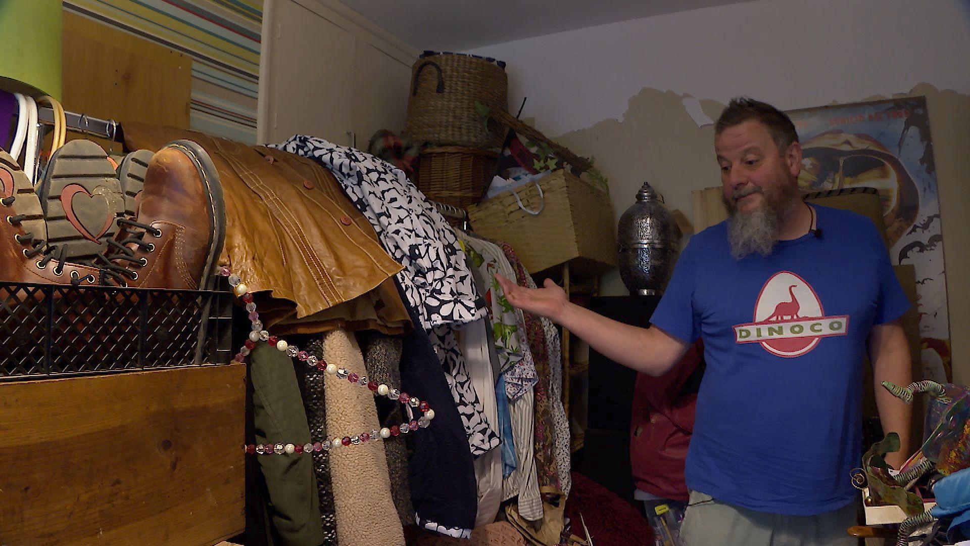 Chris Davis standing in his spare bedroom pointing to clothes that are left on rails in the middle of the room away from walls so they do not get damp.