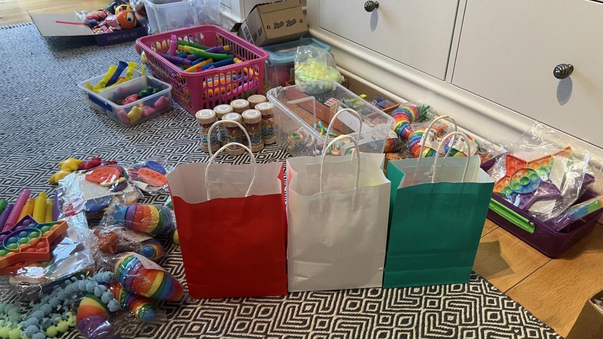 Llun o dri bag synhwyraidd Noa mewn gwahanol liwiau, coch, gwyn a gwyrdd, a rhai o'r teganau sydd yn y bagiau.