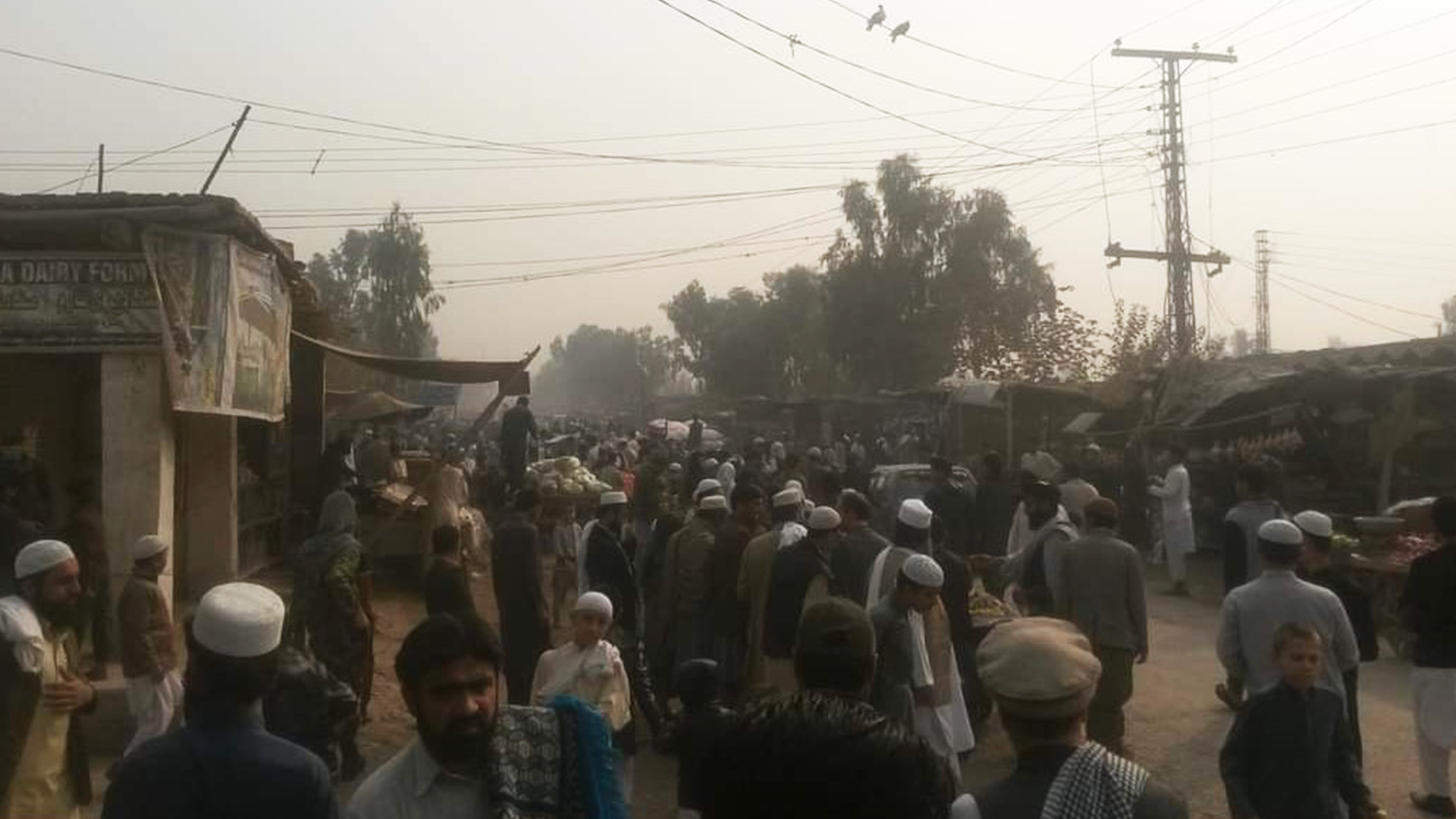 A busy, dusty street, filled with men, most of whom are wearing traditional Afghan clothing with brimless hats. There are ramshackle buildings and pylons hold up power lines across a backdrop of a grey sky.
