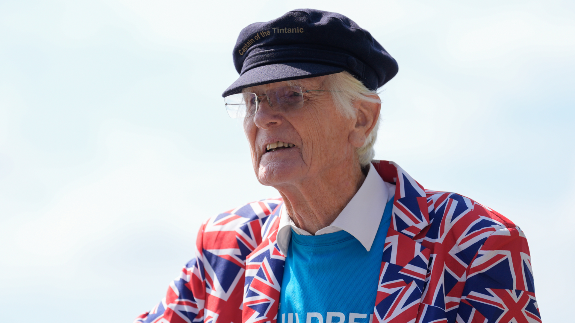 An elderly man wearing a blue captains hat, Union flag jacket, light blue jumper a white shirt. He is looking off to the left side of the image.