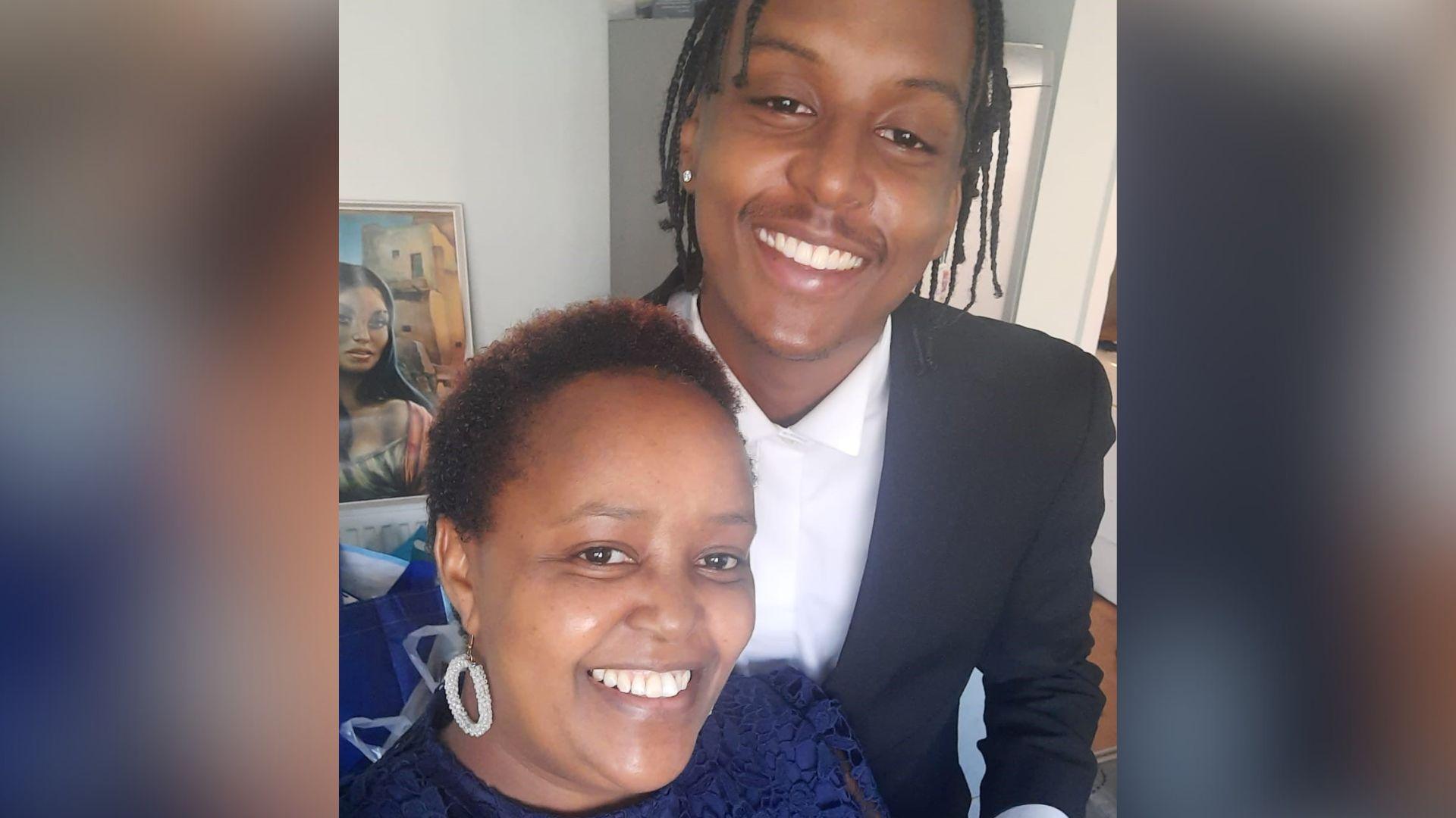 A mother and son pose for a selfie. She is standing in front of him and he is a whole head taller than her. They are both smiling broadly at the camera and are smartly dressed, with Mr Kinuthia wearing a black suit jacket and white shirt. They appear to be standing in a house with a painting in the background.