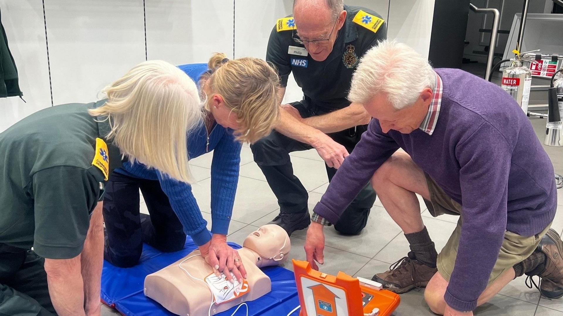 Two members of the SWAST in their bottle green uniform with the NHS logo on the front lean over an resus manikin while a man and a woman lean over the doll and the woman gives chest compressions with her hands clasped over each other.