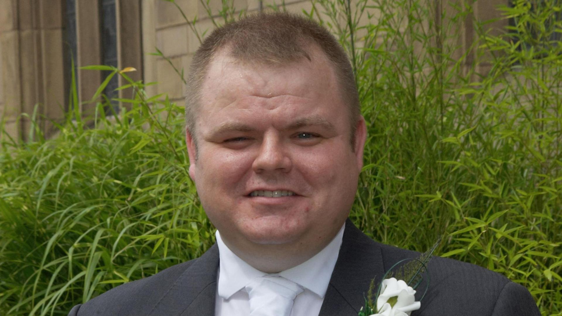 PC Neil Doyle, who has short light brown hair, smiles at the camera while wearing a grey wedding suit with a white flower attached to the lapel