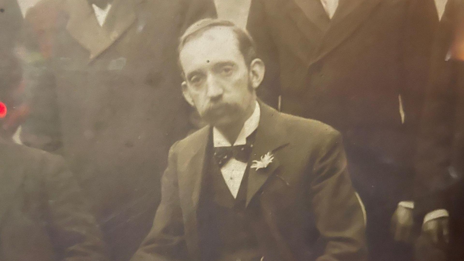 A sepia photo from the early twentieth century of a man with a large moustache  in a three-piece suit and spotted bow-tie.