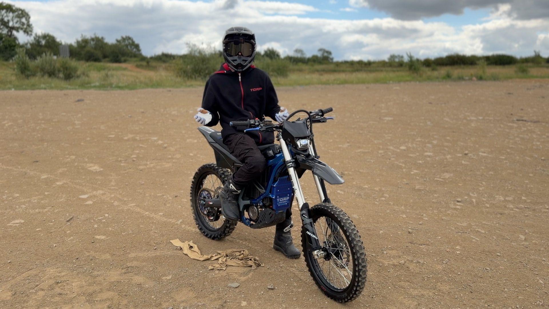 A person on a motorcycle wearing a helmet and dark clothing. They are in the middle of a piece of bare land, with a dusty gravel on the floor, and some low shrubs and trees in the background.