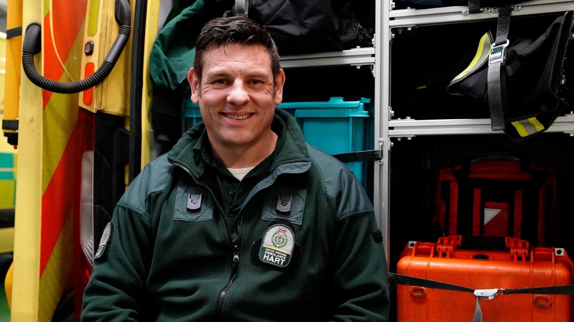 Craig is sat next to the truck containing their specialist equipment. He has short, dark hair and is wearing a green paramedic's uniform.