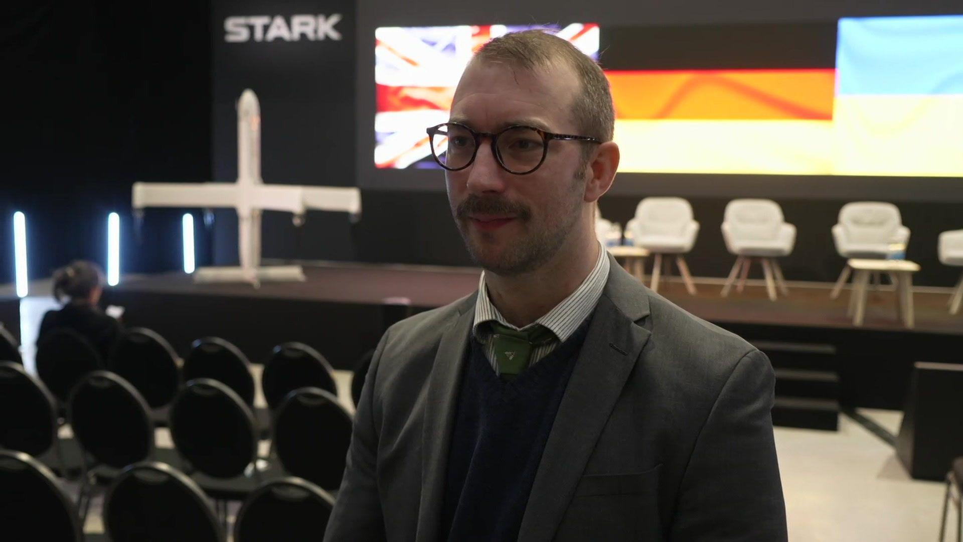 Will Stone looks to a camera slightly to the side - he is wearing glasses, a grey suit jacket with a jumper, a tie and a stripy shirt. Behind him is an empty stage, set up as if for people sitting to talk to an audience.