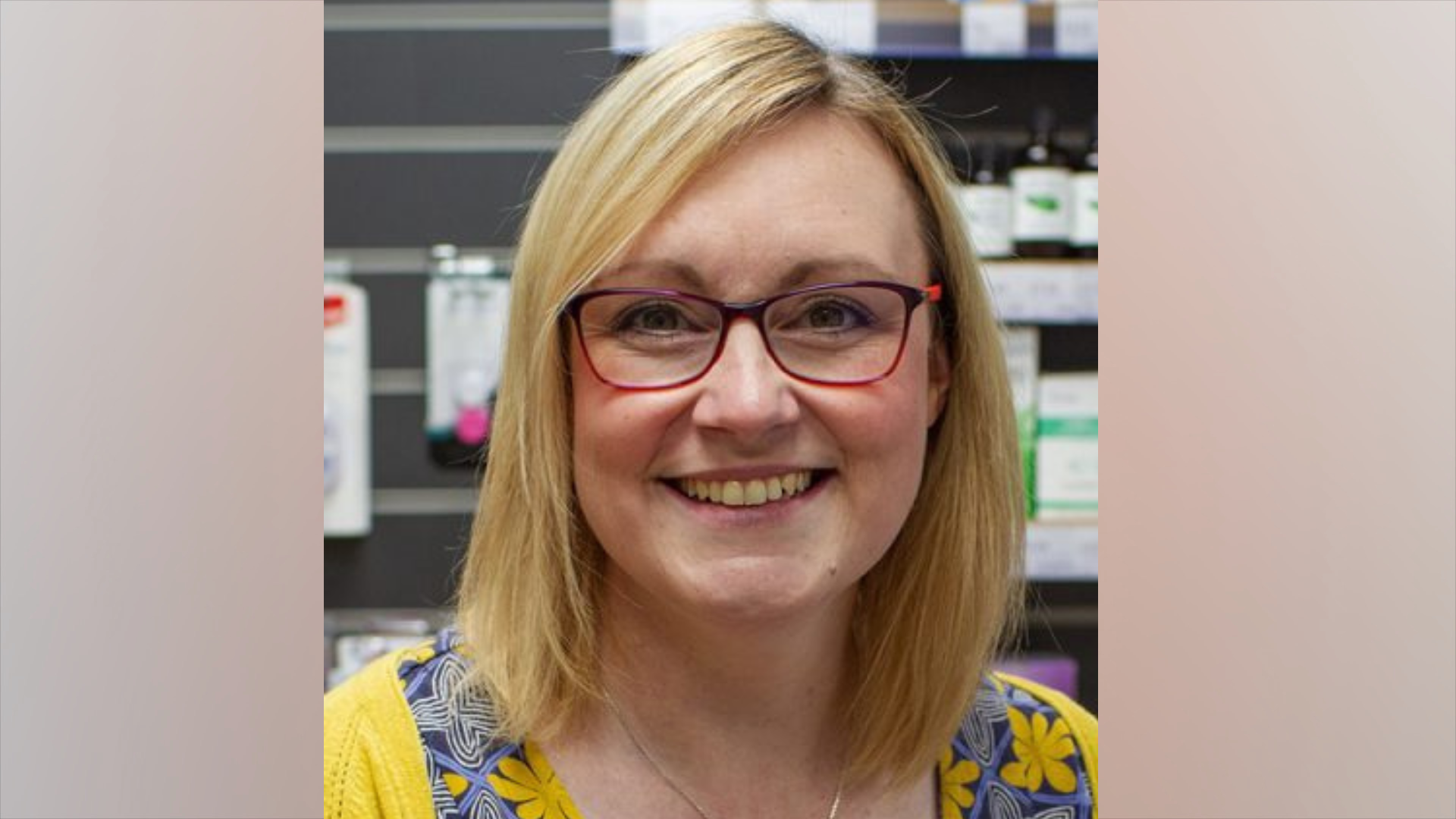 A woman with blonde hair and wearing glasses smiles at the camera, in a pharmacy.