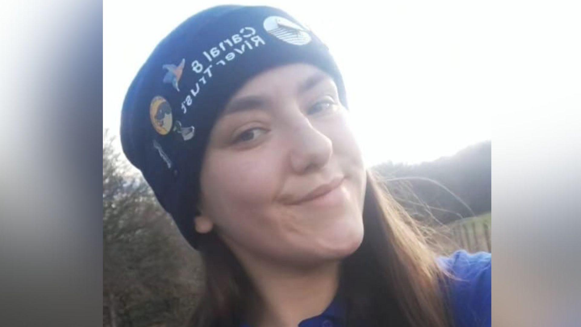 Eve wearing a dark blue hat decorated with multiple colourful embroidered patches. The individual has long brown hair and is dressed in a blue top. The background shows an outdoor setting with trees and a wooden fence.
