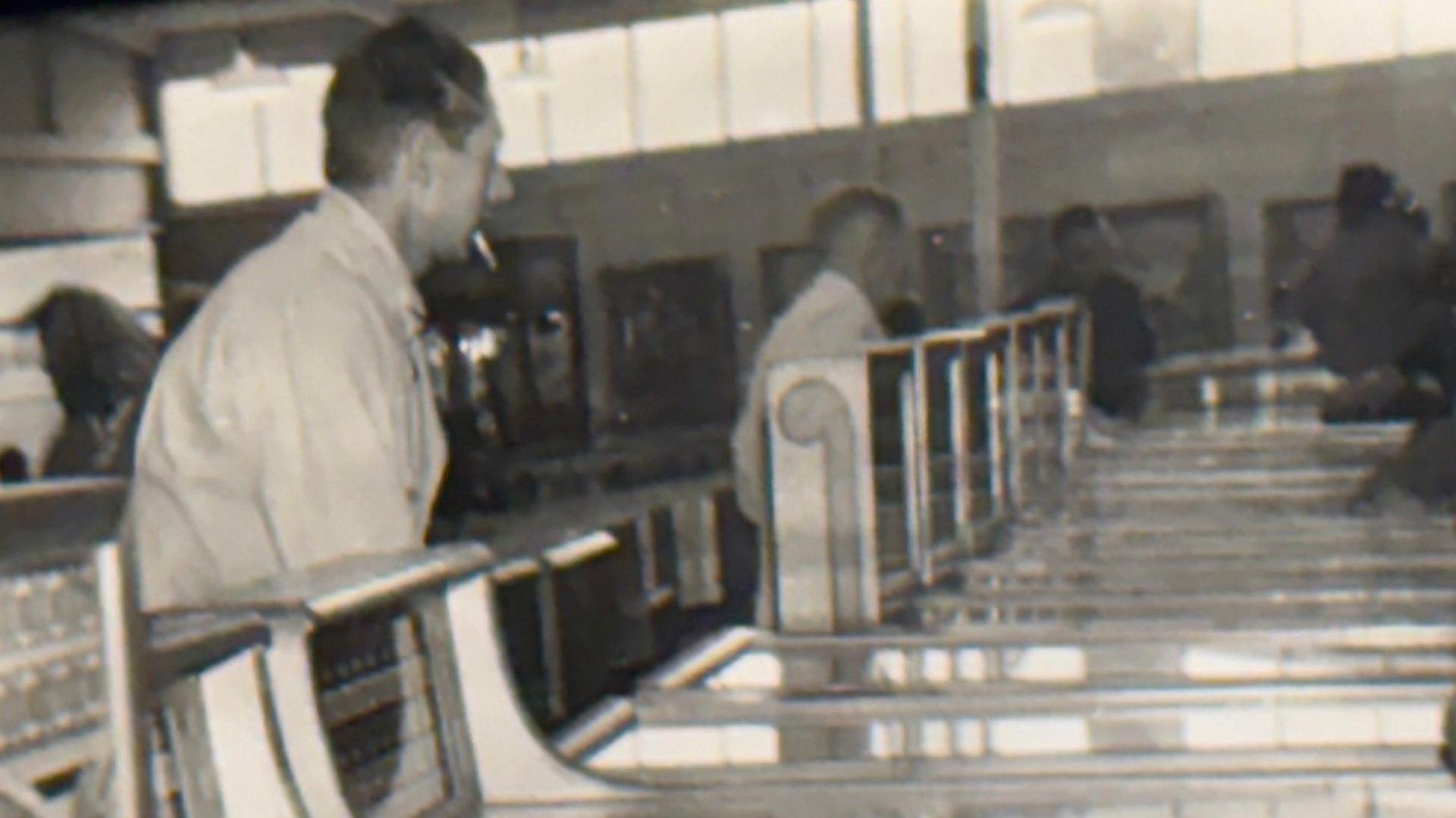 Bill Cammack (Gareth's grandfather) tends to the machines. Bill has a cigarette in his mouth. He is standing in front of a line of gaming machines.