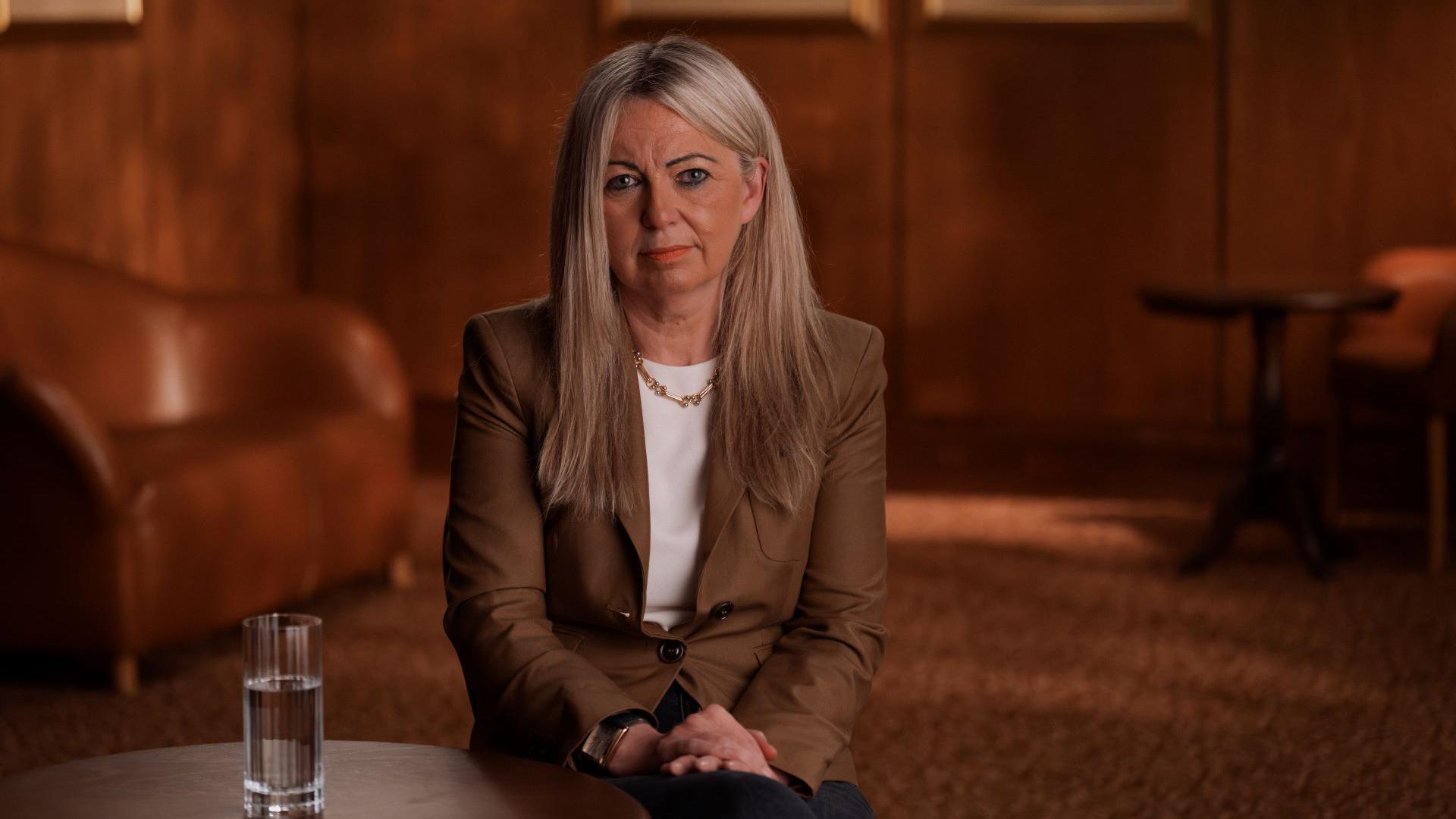 Fiona is photographed against brown background. She is looking stright at the camera. She has long blonde hair and is wearing a brown jacket with a white shirt underneath and a necklace.