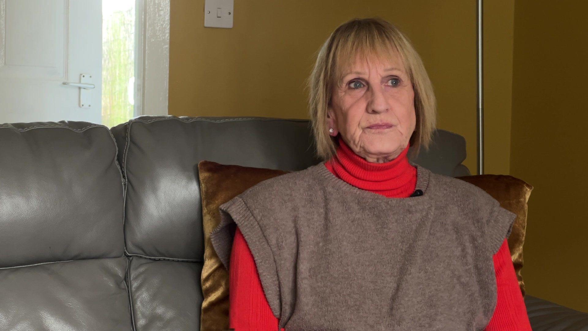 Wendy Hutchinson, sat on a grey sofa and looking away from the camera. She has medium-length blonde hair and is wearing a red turtleneck top with a grey sleeveless jumper over the top. 