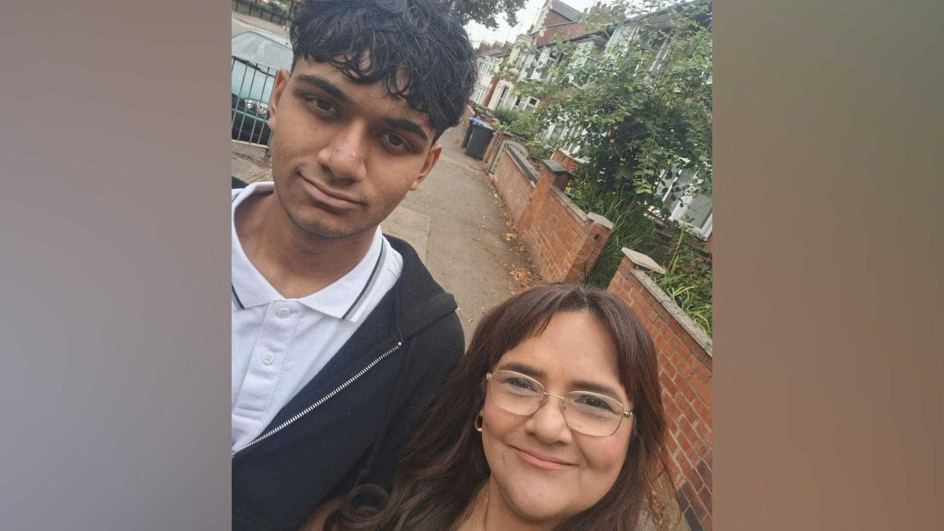 Rufia Ashraf with her son Emraan. He is to the left of the picture and has black hair. He is of Bangladeshi heritage and is wearing a white polo shirt with black edging and a black hoodie. Rufia has brown hair and glasses. She is wearing hooped earrings. Her fringe is parted towards the middle.