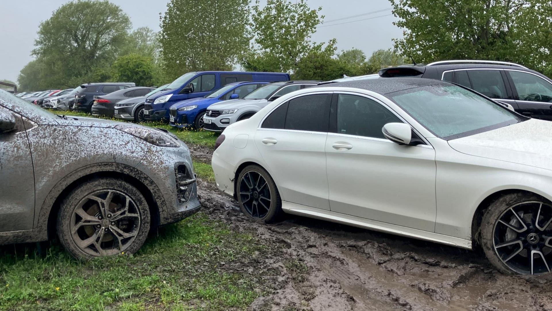 Multiple cars are parked on a muddy field. One white car is parked in a very muddy patch and a silver car has dried mud which had sprayed from another car all over it.