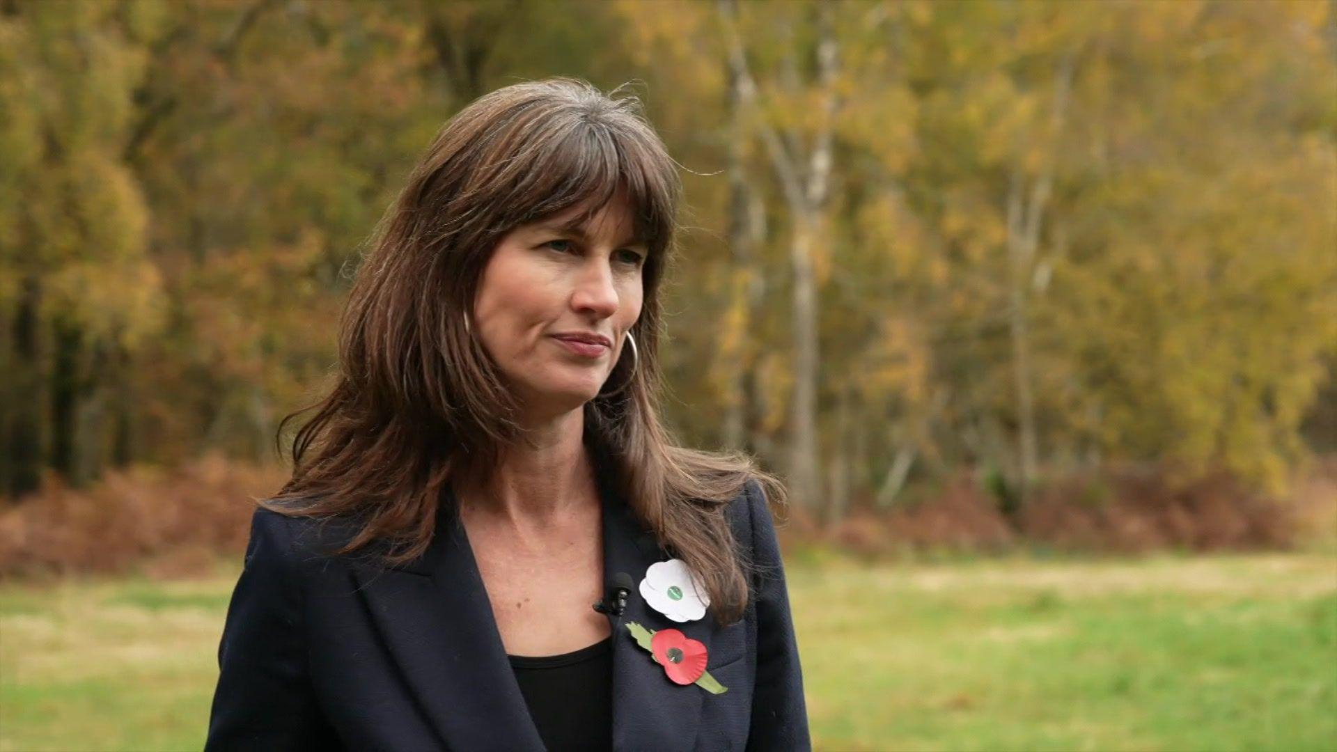 A woman with brown hair and a fringe. She is wearing a black blazer with a red poppy on it.