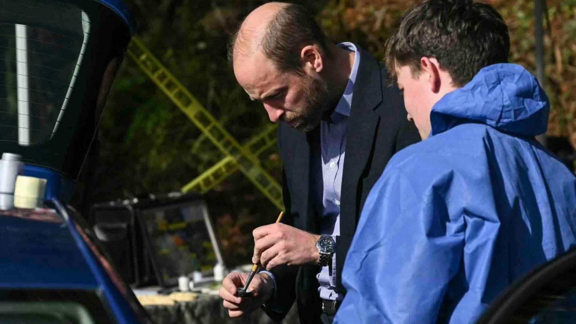 The Prince of Wales in a dark jacket and blue shirt holds a brush in his hand with a young person in a blue overall looking at him with the side of a car on the left.