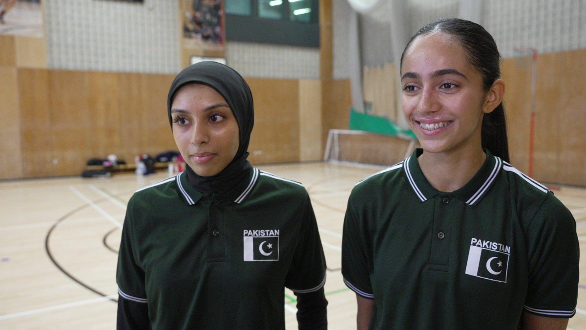 Leya Shah stands on the left looking off from the camera. She wears a black hijab and black polo top with a Pakistan logo top left. Sumayya is on her right, also looking past the camera and smiling widely. Her hair is pulled back in a ponytail and she is also wearing a black polo top with a Pakistan team logo top left