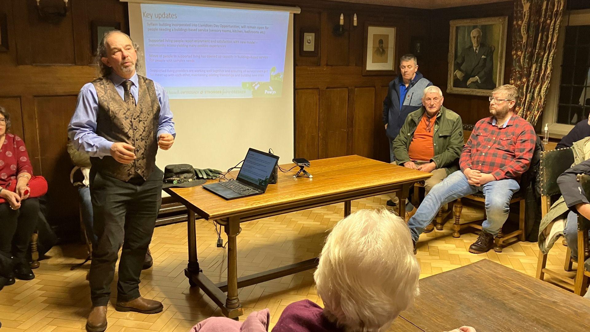 Llun o'r Cynghorydd Pete Roberts yn siarad o flaen pobl mewn cyfarfod. Mae powerpoint ymlaen ar y wal y tu ôl iddo a gliniadur ar fwrdd wrth ei ymyl.