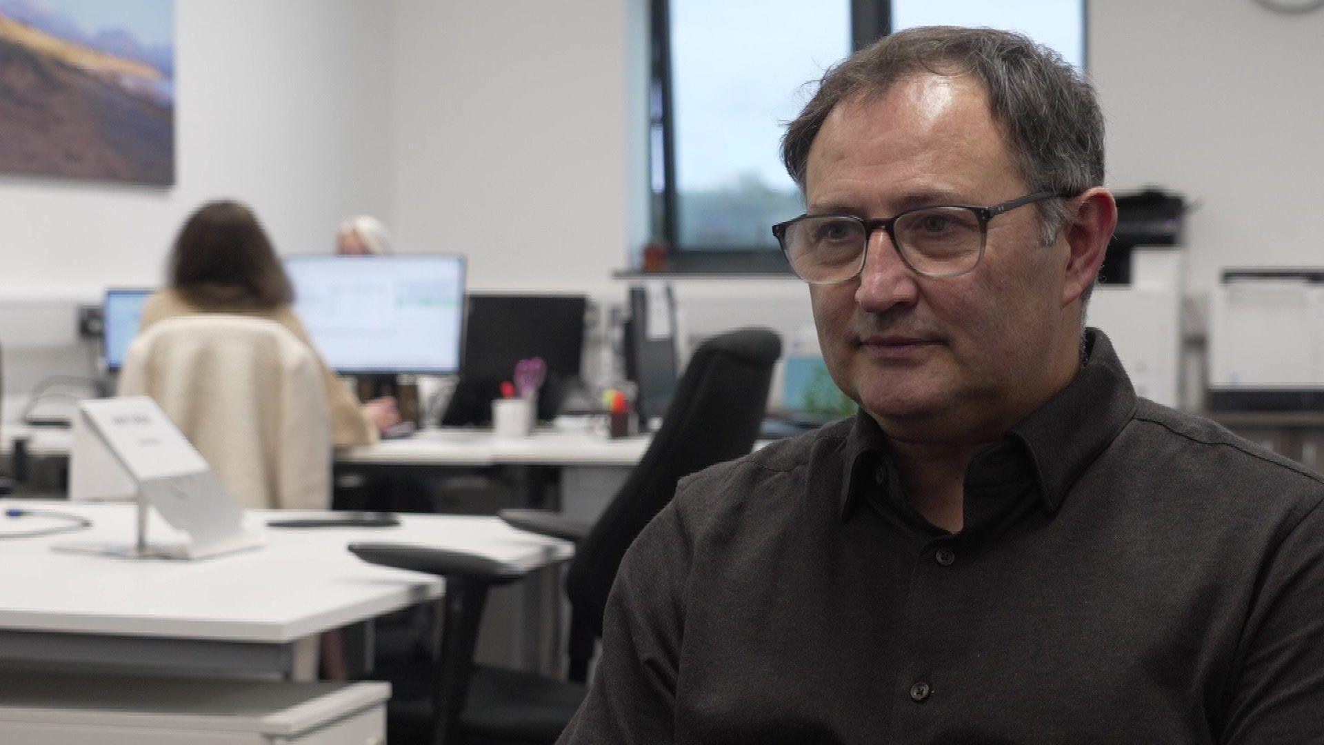 Huw Watkins, dyn mewn sbectol a chrys tywyll, yn eistedd yn ei swyddfa