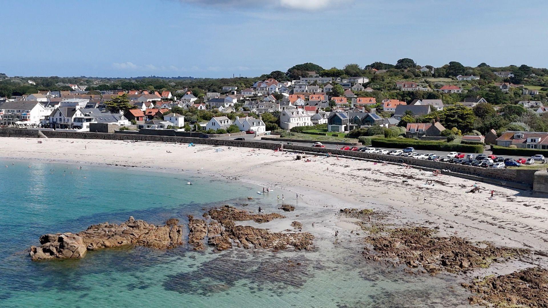 A sandy beach with a rocky outcrop and sea defences, a car park and houses behind.