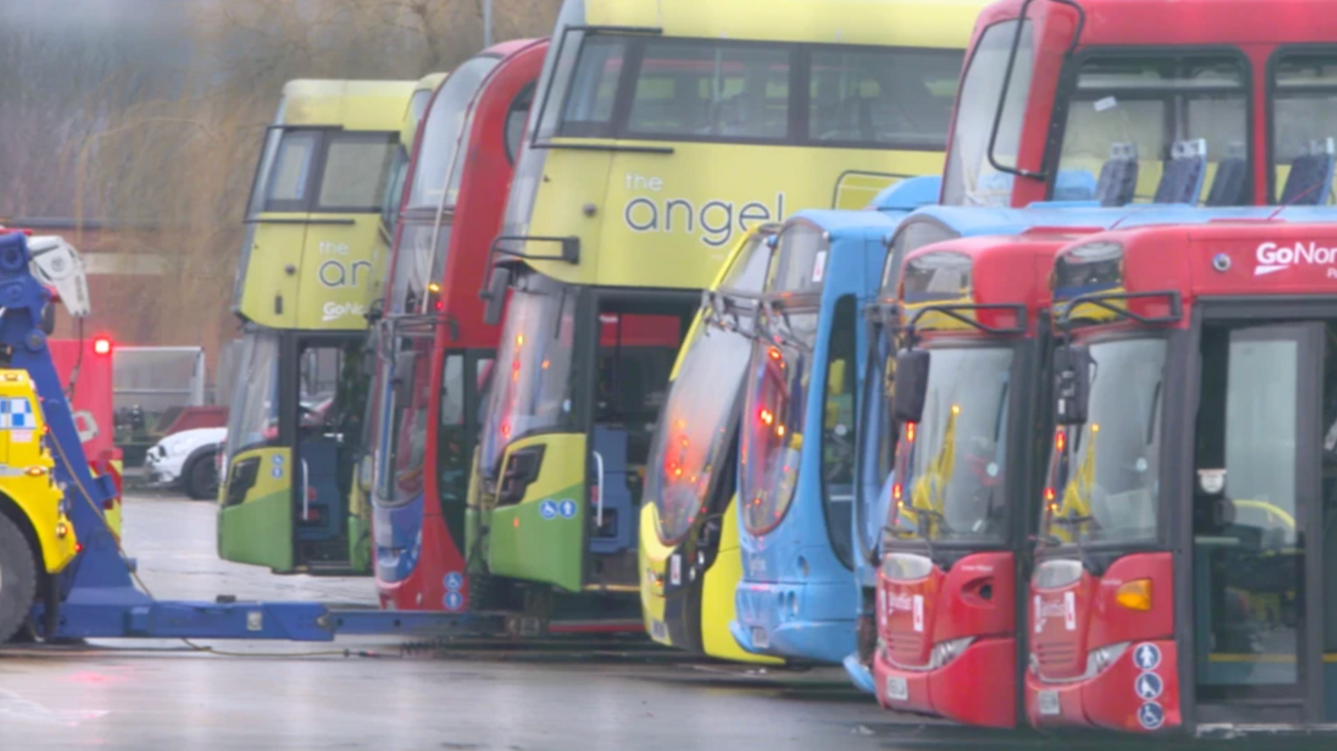 A yellow recovery truck is attached to a yellow and green double-decker bus which says 'Angel' on the side. It is surrounded by other buses which are off the road.