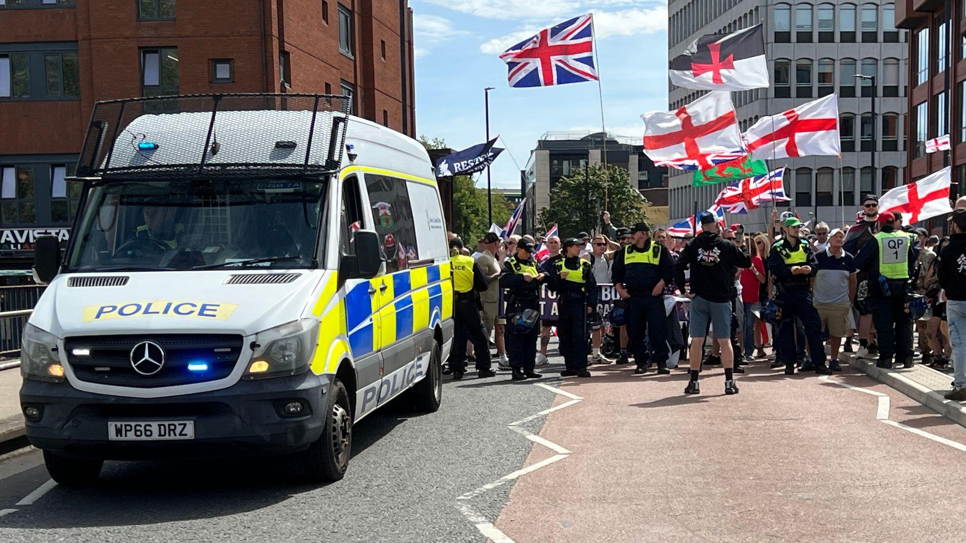 A police riot van parked on a bridge, which is blocked by protesters standing behind a line of police officers. The group hold flags, including Union and St Gorge flags and the Welsh flag. 