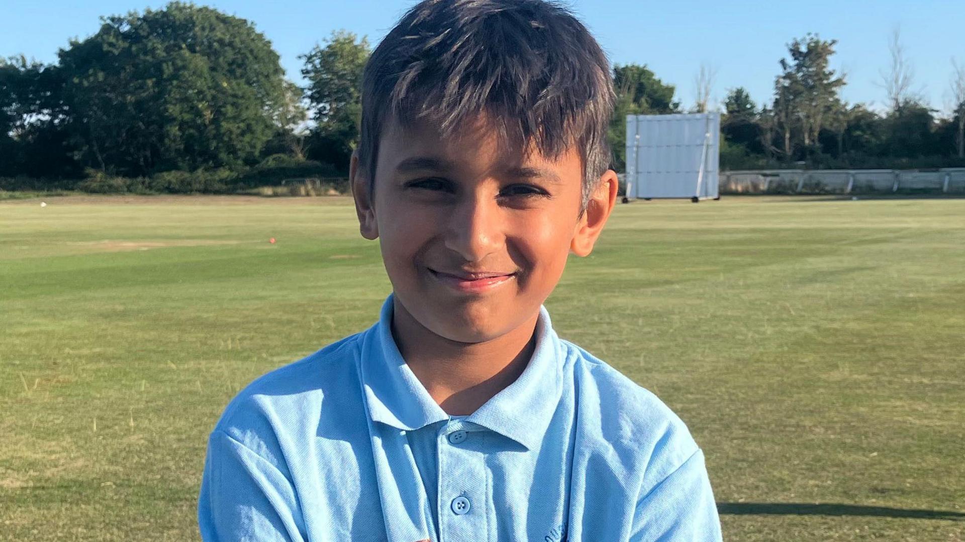 Om is standing on the short green grass of the cricket grounds and smiling at the camera on a sunny day with blue skies. He has short dark hair and is wearing a light blue cricket shirt with a collar buttoned up.