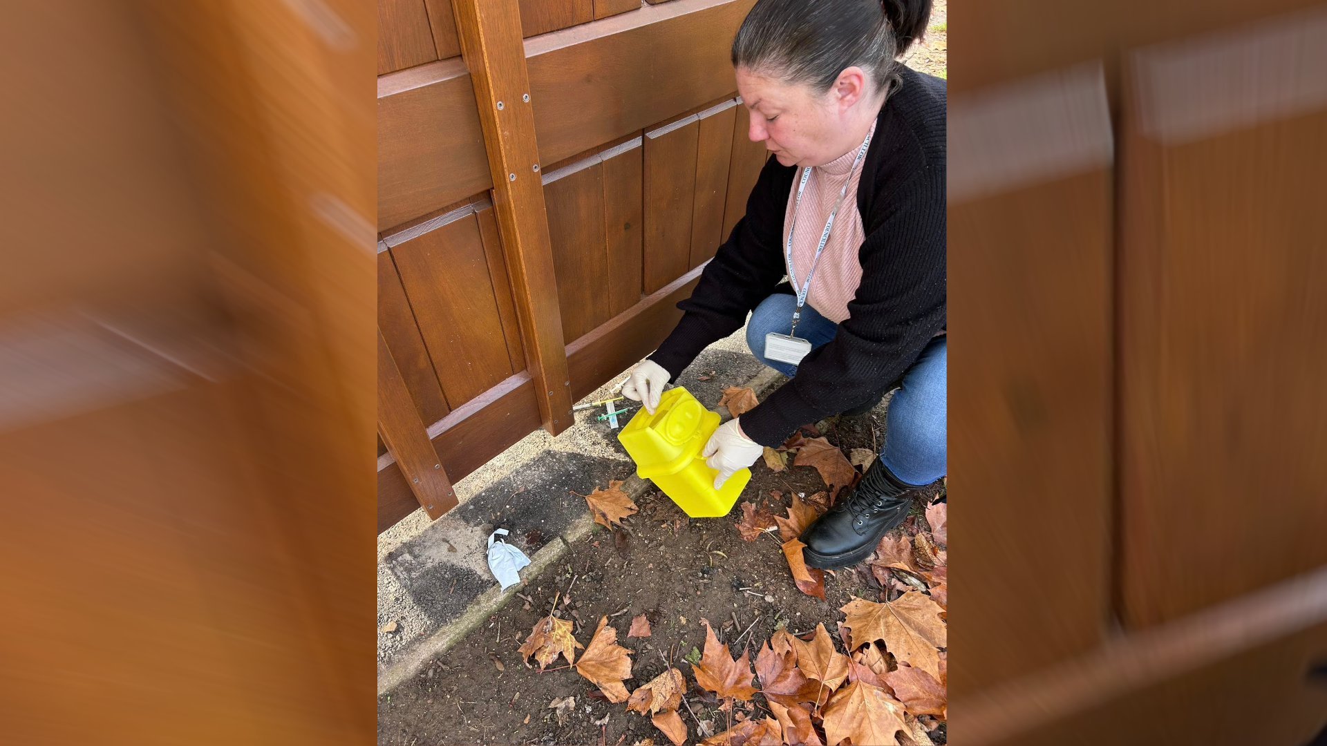 Kirsty, a woman wearing with brown hair, wearing a black cardigan and a pink shirt- holding a yellow needle box collecting needles.