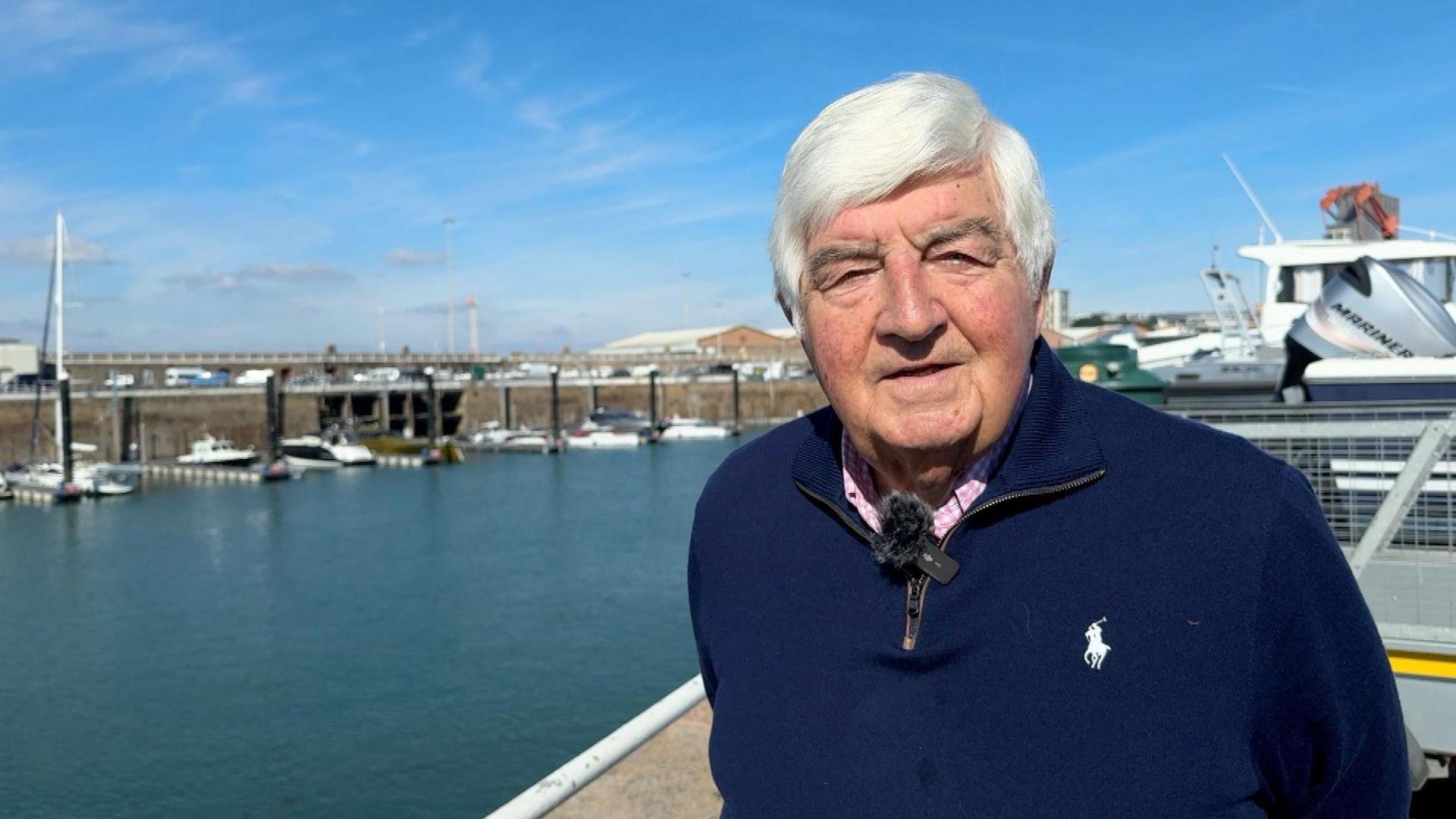 Brian Nibbs - a gentleman with white hair wearing a branded navy zip up jumper, with a white and pink checkered collared shirt underneath. Behind him is the harbour with various vessels docked in the distance.