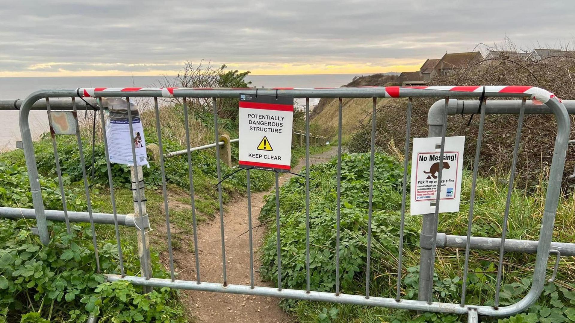 A metal barrier is across a pedestrian access point to a beach. A sign on the barrier says: "Potentially dangerous structure - keep clear." Steps down the cliff can be seen in the background and the roofs of houses near the edge of the cliff are also visible.