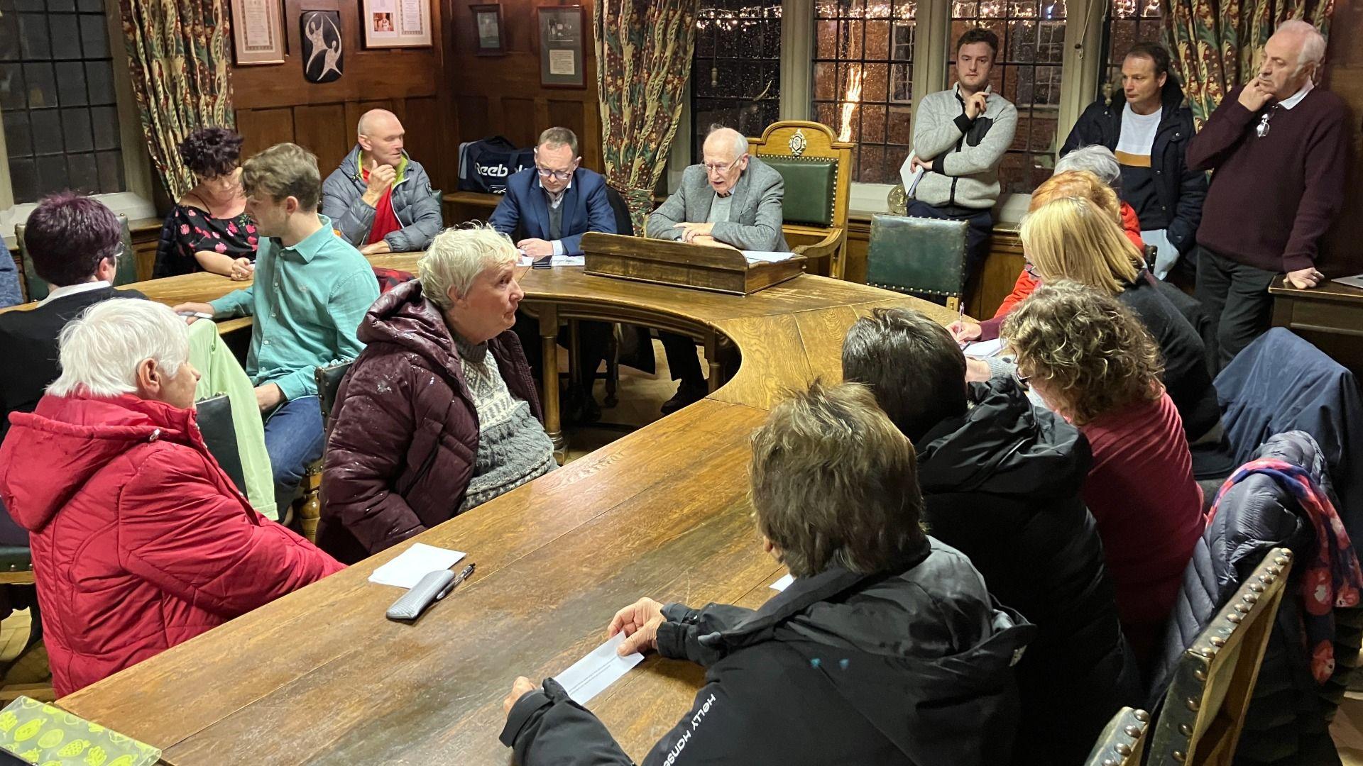 Llun o bobl yn eistedd o gwmpas bwrdd hir a chrwn.