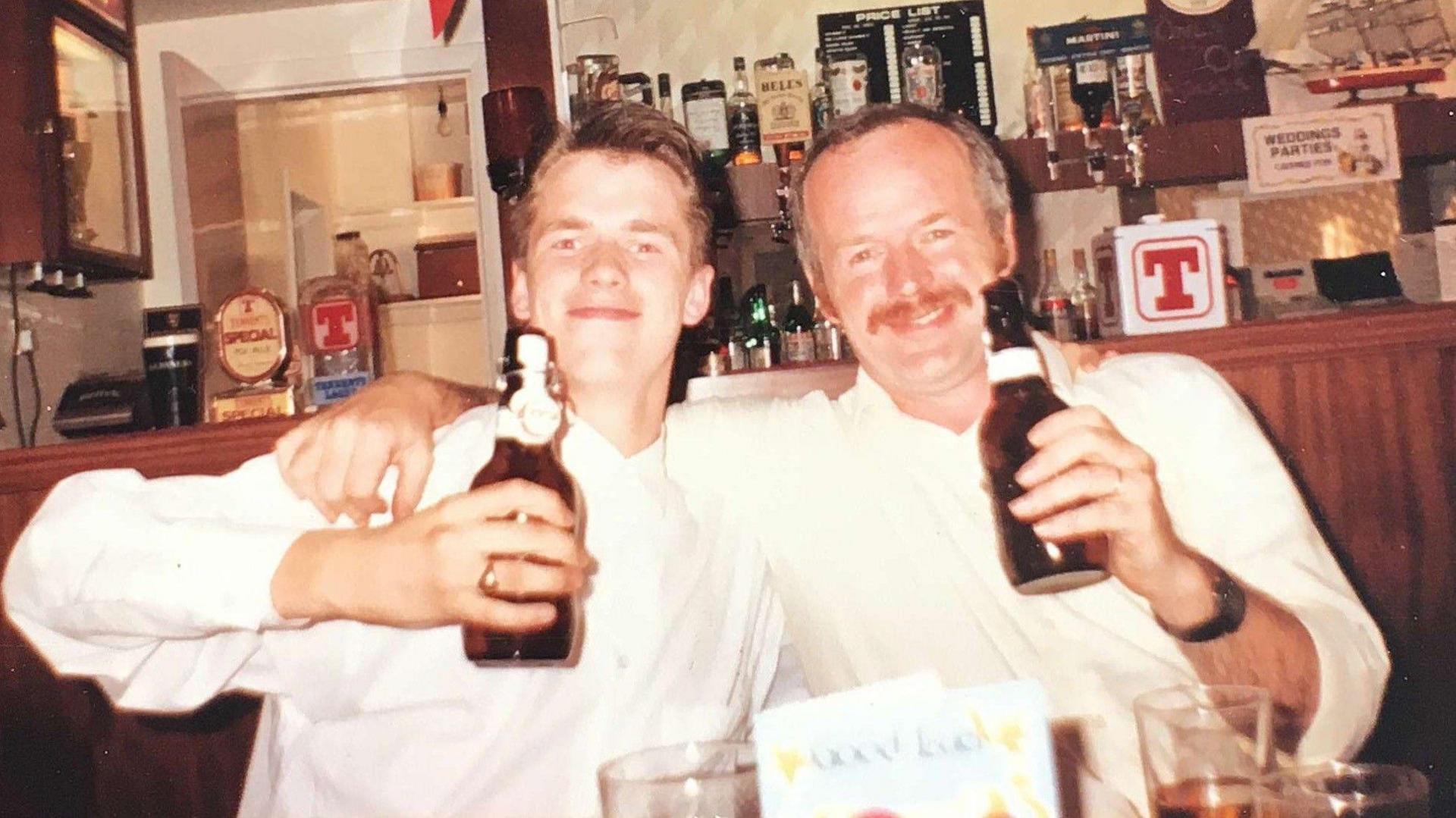 Shane and David Gorman, wearing similar white shirts, their arms around one another, toast the camera with bottles of beer in a bar.