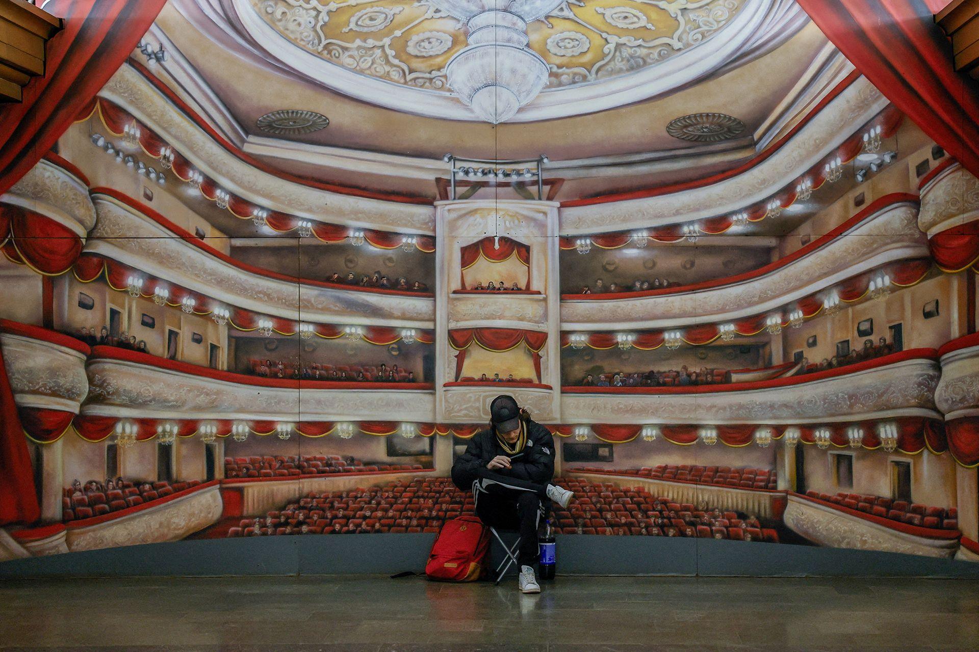 A person sits cross-legged in a metro station in Kyiv, in front of a mural depicting a large theatre, during an air raid in Kyiv in March 2024.